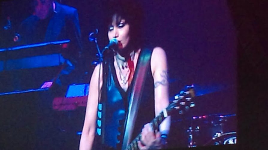 Joan Jett performing live onstage, singing into a microphone while playing electric guitar under blue and purple concert lighting.