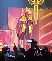 Rob Halford of Judas Priest performing onstage in a shiny, embellished jacket under colorful concert lighting.