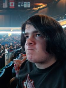 The Kid sitting at a Judas Priest concert with an unimpressed expression as the crowd gathers behind him.
