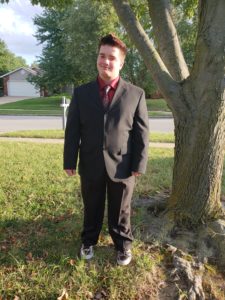 The Kid dressed for Homecoming in a dark suit, red shirt, and tie, standing on the grass beside a tree.