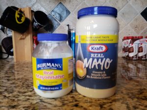 Two jars of mayonnaise on a kitchen counter, with an oversized jar of Kraft Real Mayo towering over a smaller jar of Burman's mayonnaise.