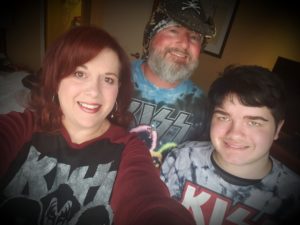 Kat, The Husband Dude, and The Kid smiling together in a family selfie, all wearing KISS band shirts.