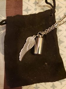 Silver memorial necklace featuring an angel wing charm and a small cylindrical pendant containing E's ashes, resting on a black drawstring pouch.