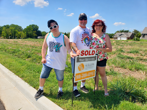 Kat stands with the Husband Dude and the Kid on a sunny, undeveloped lot, celebrating the purchase of their future homesite. The family poses behind a real estate sign marked “SOLD.”